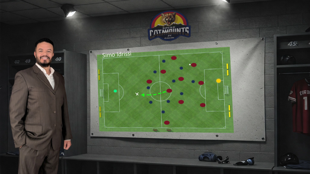 A man (Simo Idrissi) in a suit is standing in a locker room. On the wall behind him, there is a large tactical board displaying a soccer field with player positions marked by red and blue dots. The board also includes strategic markings, such as a green arrow indicating a movement or pass. Above the board, there is a logo for 'Rocky Catamounts FC Colorado.' The name 'Simo Idrissi' is written on the top left corner of the board. The locker room has numbered lockers, and there is a jersey with the number 1 hanging on the right side. The image highlights a strategic planning session for a soccer team, emphasizing the tactical aspects of the sport.