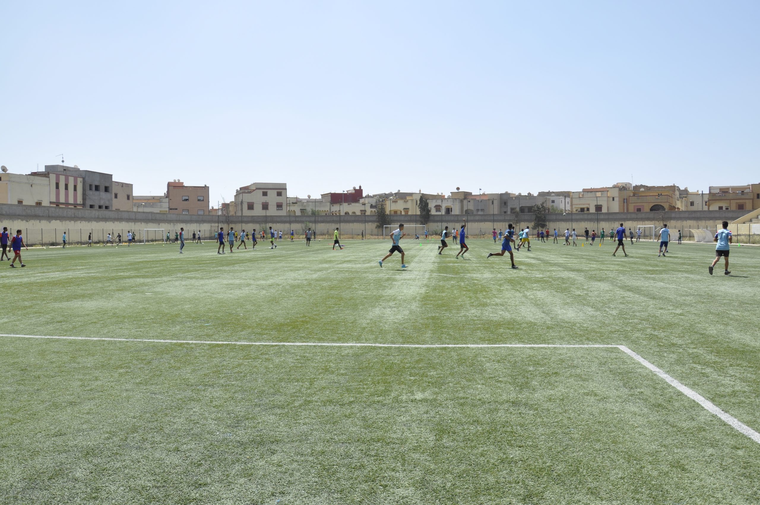 Simo Idrissi a Colorado Leader with Soccer Coaching Philosophy. Adult male soccer players train on a large artificial turf field enclosed by a tall metal fence in an urban setting. The session is led by Colorado coach Simo Idrissi. A total of 16 players are visible: 7 in red jerseys, 6 in black, and 3 in white. The players are spread across the field in active formations, suggesting structured drills or small-sided games. Residential buildings with flat roofs and mixed architectural styles line the background under a clear, sunny sky.