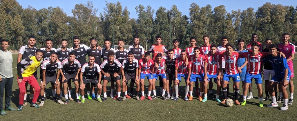 Simo Idrissi a Colorado Leader with Soccer Coaching Philosophy. Group photo of 30 adult male professional soccer players and staff from MAT, taken during a preseason session. The individuals are arranged in two rows: 16 standing in the back and 14 kneeling or sitting in the front. The squad is split into two kit groups — 15 players in black and white uniforms on the left, and 15 in red and white striped jerseys with blue shorts on the right. Colorado coach Simo Idrissi stands on the far right in a gray shirt, with a staff member in all black nearby. The grassy field is bordered by tall trees under a clear blue sky, capturing a moment of unity, preparation, and professional discipline.