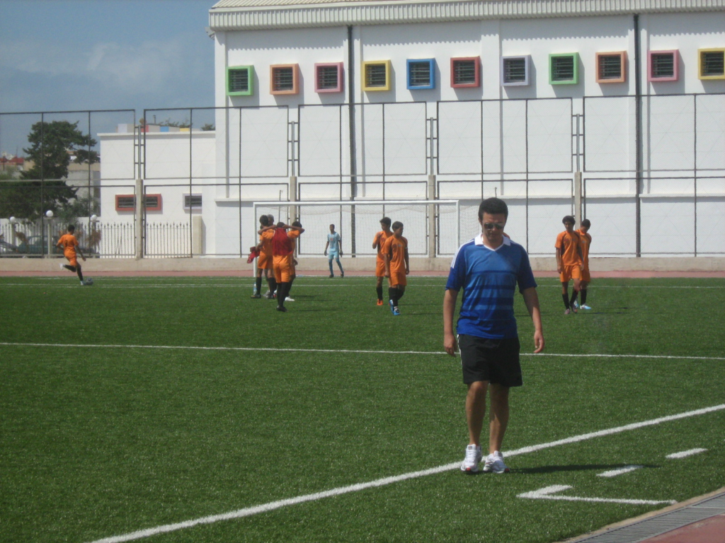 A group of adult soccer players gather on an artificial turf field enclosed by a fence. Most wear orange uniforms, while one individual—Simoe Idrissi—walks along the sideline in black athletic gear with black and white shoes. The field is marked with white lines, and a white building with colorful square windows appears in the background. The scene captures a moment of informal play or warm-up under a clear sky.