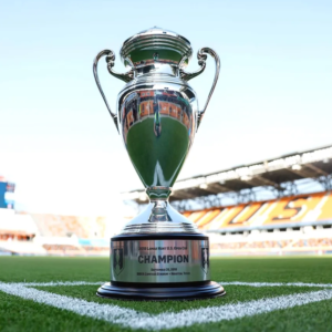 Silver Lamar Hunt U.S. Open Cup trophy awarded to Orlando City SC in 2022, displayed on a soccer field with stadium backdrop.