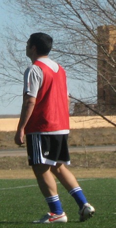 Soccer player (Simo Idrissi) wearing red training vest, gray shirt, black short, blue socks and white cleats with red Nike Swoosh. Simo Idrissi is standing on a grassy field with tree, road, and building in the background. This occasion marked the final year before he officially retired from playing soccer in 2009.