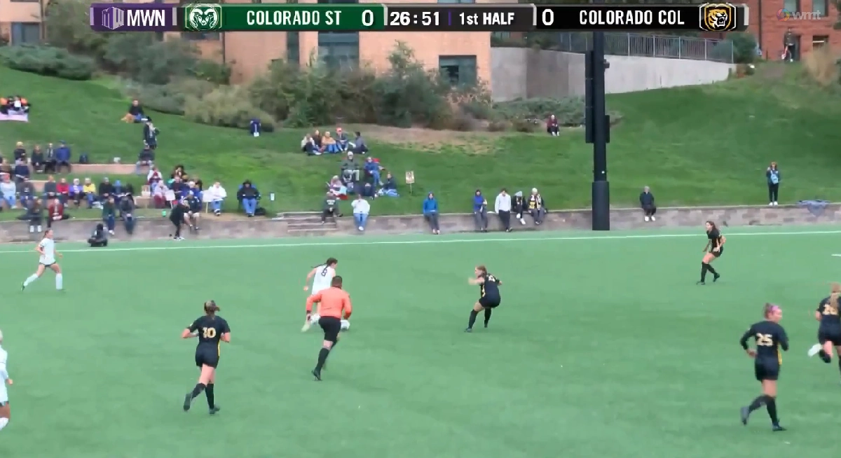 Colorado State and Colorado Boulder women’s soccer teams compete on a green turf field, with the scoreboard showing a 0–0 tie in the first half at 26:51. Colorado State players wear white uniforms, while Colorado Boulder athletes are in black with gold numbers. Spectators watch from a grassy hillside in the background.