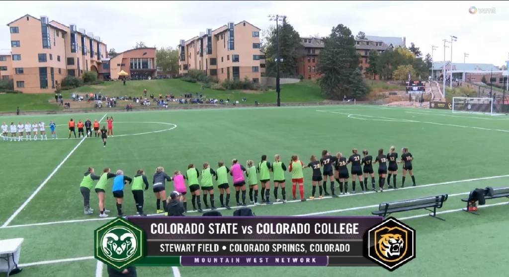 Players from Colorado College and Colorado State line up on Stewart Field in Colorado Springs for a women’s soccer match, broadcast by the Mountain West Network. Colorado College athletes wear black uniforms, while Colorado State players sport green pinnies. The artificial turf field is framed by residential buildings and spectators seated on the grassy hillside.